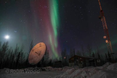 Earth's originally satellite the moon, a satellite receiving dish and radio tower, all visible on top of Ski Boot Hill as northern lights streak overhead. 